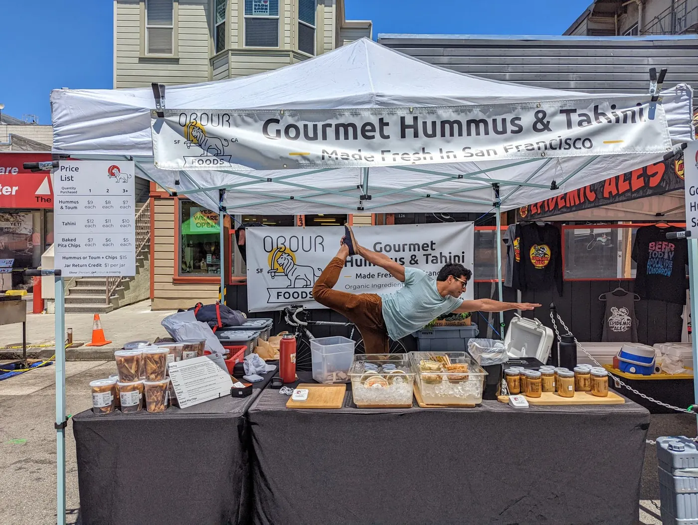 The Obour stand at a San Francisco street market: a white canopy with Obour banners, a table of hummus, toum, tahini and pita chips, and a team member striking a yoga pose behind it